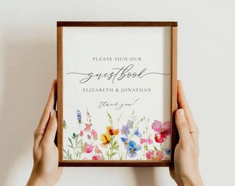 May include: A wooden framed guestbook sign with floral watercolor art. The sign reads "Please sign our guestbook" with the names "Elizabeth & Jonathan" and "Thank you!" in elegant script.