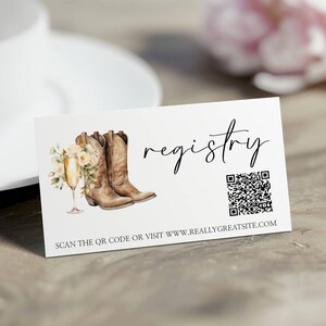K&ouml;nnte beinhalten: Wei&szlig;e Karte mit dem Wort &bdquo;registry&ldquo; in Schreibschrift. Die Karte zeigt eine Aquarellillustration von braunen Cowboy-Stiefeln, einem Sektglas und Blumen. Ein QR-Code und eine Website-Adresse sind ebenfalls enthalten.