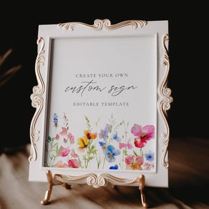May include: A white framed sign with gold accents, displaying the text "CREATE YOUR OWN custom sign EDITABLE TEMPLATE." The sign features a watercolor floral design with pink, yellow, and blue flowers. It is propped up on a gold easel.