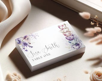 May include: A white place card with floral and teacup illustrations. The card features the name "Lisa Smith" in cursive, with "TABLE ONE" below. Purple and lavender flowers and teacups decorate the card.