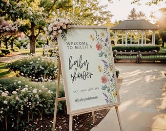 May include: A wooden easel sign with a floral arrangement and the words "Welcome to Millie's baby shower." The sign is decorated with colorful flowers and greenery. The event details are also included on the sign.