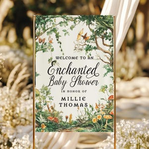 May include: A rectangular welcome sign for a baby shower featuring a cream background with a watercolor forest illustration of green trees, colorful butterflies, and mushrooms. The text reads: Welcome to an Enchanted Baby Shower in honor of Millie Thomas.