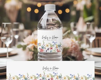 Könnte beinhalten: Eine transparente Plastikwasserflasche mit einem weißen Etikett mit dem Text "baby in bloom" und floralen Illustrationen in Pastellfarben. Die Flasche hat einen weißen Deckel und steht auf einem Tisch mit Partydekorationen.