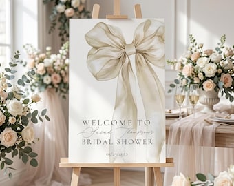 May include: A welcome sign for a bridal shower on a wooden easel. The sign displays a large, elegant bow in beige and cream tones. The text reads "Welcome to Sarah Thompson's Bridal Shower" and the date.