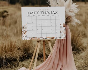 Può includere: Un cartello bianco con la scritta "BABY THOMAS" e una griglia di calendario, esposto su un cavalletto di legno. Il cartello è decorato con illustrazioni floreali nei toni del marrone e del pesca. Un tessuto rosa drappeggia sotto, con erbe alte e secche sullo sfondo.