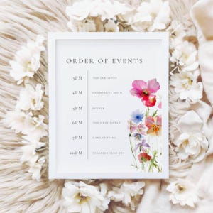 以下が含まれることがあります： 「ORDER OF EVENTS」の文字と結婚式のスケジュールが書かれた白いフレームのサイン。スケジュールには、セレモニー、シャンパンアワー、ディナー、ファーストダンス、ケーキカット、スパークラーの打ち上げの時間が含まれています。水彩画のフローラルアートが右側に飾られています。