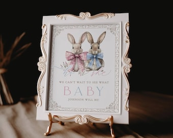 May include: A framed baby announcement features two watercolor bunnies wearing bow ties. The text reads "He or She" and "We can't wait to see what BABY Johnson will be!" The frame is white with gold accents, and the announcement is displayed on a gold easel.