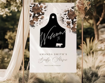 May include: A white welcome sign with brown watercolour splatters. The sign features a black silhouette of a cow tag with the word "Welcome" written in cursive. The text "Amanda Smith's Baby Shower #HolyCowItsABaby" is printed below the tag.