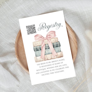 Puede incluir: Una tarjeta blanca con un lazo rosa y libros apilados. La tarjeta dice "Registry" e incluye un código QR. El texto dice "The bride-to-be, (bride's name) is registered at (name of boutique), wishing her joy, love, and laughter as we celebrate this exciting time together!"