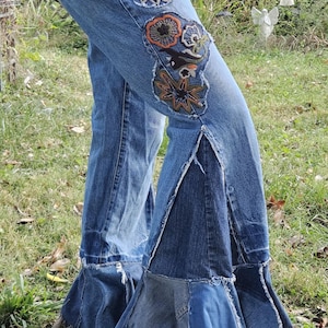 May include: Light blue denim jeans with floral embroidery on the thighs and flared bottoms. The jeans have a split seam detail at the knee and a frayed hem. The embroidery features orange, green, and black accents.
