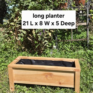 May include: A rectangular wooden planter with a natural light brown finish and a black interior liner. The planter features four short legs and is labeled with the text long planter 21 L x 8 W x 5 Deep, indicating its dimensions in inches.