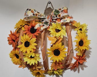 May include: A brown and orange pumpkin-shaped wreath decorated with sunflowers and fall leaves. The wreath is tied with a white and orange ribbon with a pumpkin print.