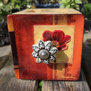 May include: A square wooden box with a vibrant orange and red finish featuring a decorative floral pattern. The front panel displays a painted red flower and a white ceramic knob with a black leaf design centered over the floral artwork.