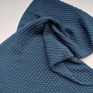 May include: A dark blue knitted blanket featuring a consistent textured honeycomb pattern throughout. The fabric is folded to show the depth of the weave. The text "Durchgehend mit muster ohne Namen" is printed in black in the upper right corner.