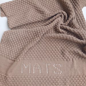 May include: A light brown knitted blanket featuring a textured diamond pattern throughout the fabric. The name MATS is knitted into the lower section in a lighter shade. The text mit Namen is printed in black at the bottom of the image.