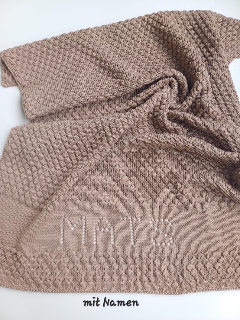 May include: A light brown knitted blanket featuring a textured diamond pattern throughout the fabric. The name MATS is knitted into the lower section in a lighter shade. The text mit Namen is printed in black at the bottom of the image.