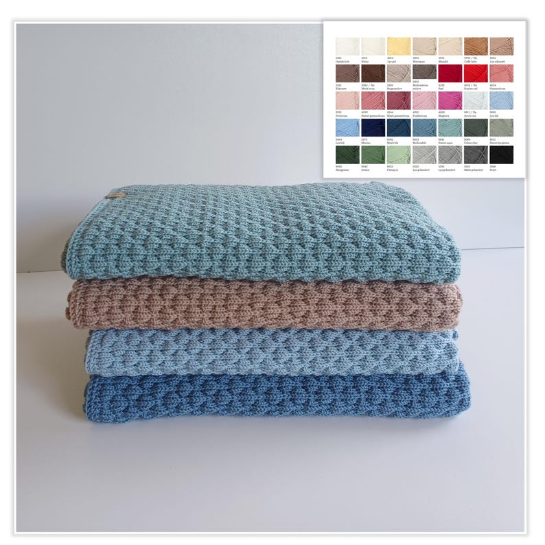 May include: A stack of four folded knitted blankets featuring a textured honeycomb pattern. The blankets are colored in shades of sage green, dusty rose, light blue, and navy blue. A color chart with various yarn swatches is visible in the corner.