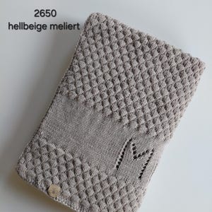 May include: A light beige heathered knit blanket featuring a textured bobble stitch pattern on the top and bottom sections, a smooth center band, and a letter M integrated into the knit. A small square tag with a bee icon is attached.