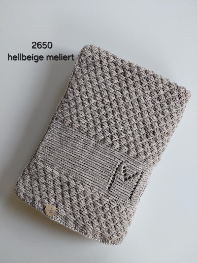 May include: A light beige heathered knit blanket featuring a textured bobble stitch pattern on the top and bottom sections, a smooth center band, and a letter M integrated into the knit. A small square tag with a bee icon is attached.
