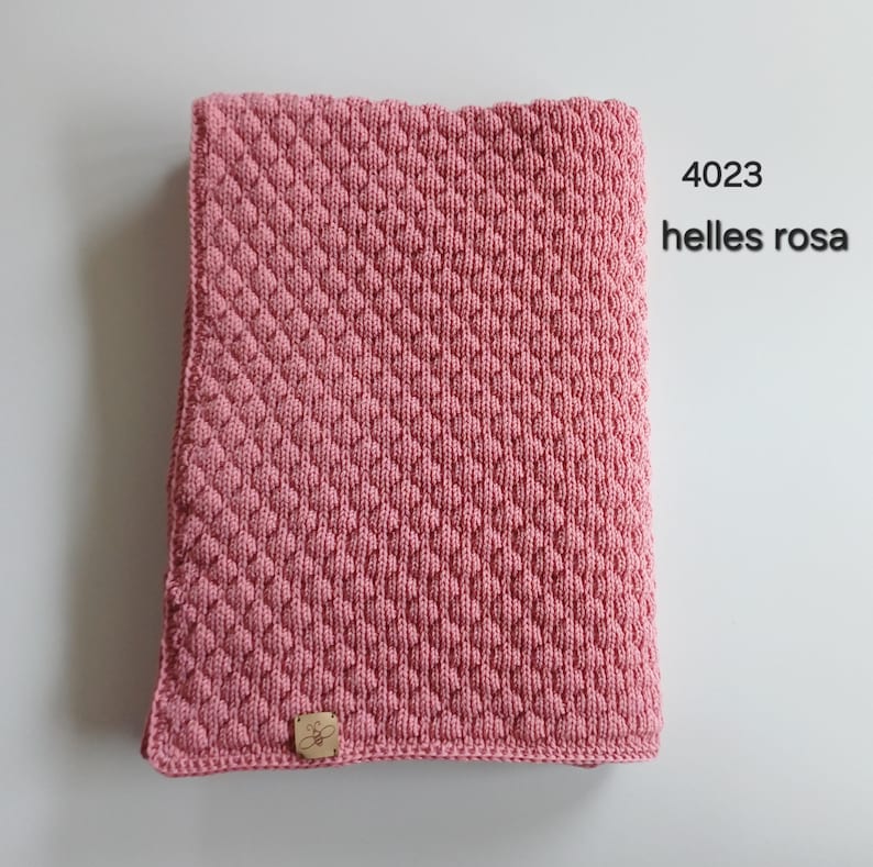 May include: A rectangular knitted blanket in a dusty pink color featuring a raised diamond texture pattern. A small square tan label with a bee icon is attached to the bottom left corner. The text 4023 helles rosa is printed in black.