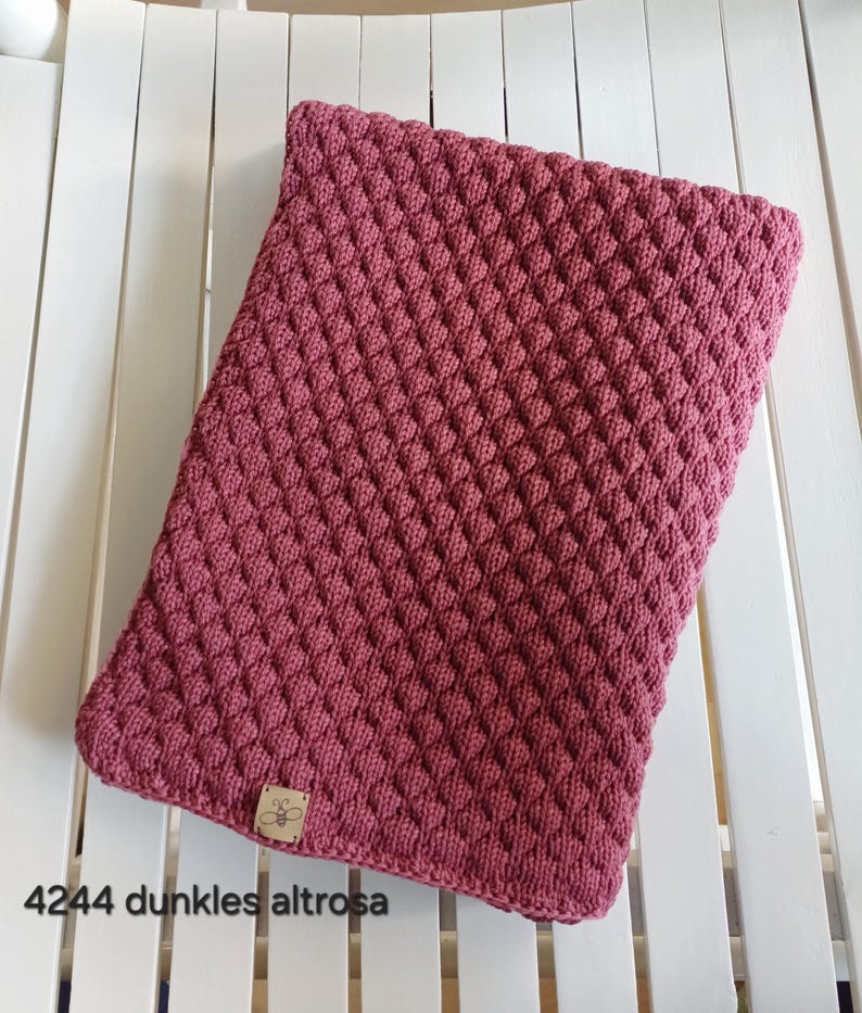 May include: A rectangular knitted blanket in a deep dusty rose color featuring a textured diamond pattern. A small square tan leather tag with a bee icon is attached to the bottom left corner. The text 4244 dunkles altrosa is visible.
