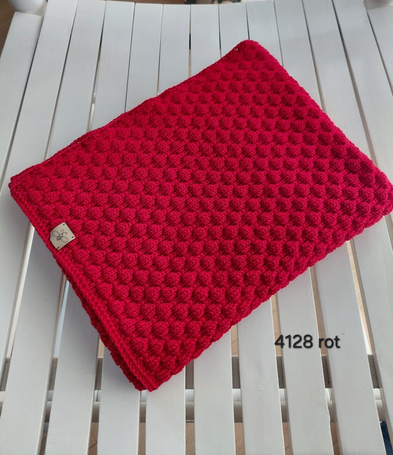 May include: A vibrant red crocheted blanket featuring a textured bobble stitch pattern throughout. A small rectangular wooden tag with a bee icon is attached to the corner. The text 4128 rot is printed in the lower right corner of the image.