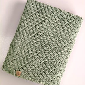May include: A rectangular sage green knitted cover featuring a textured diamond pattern. A small tan leather-like tag with a bee icon is attached to the bottom left corner. The word "pistazie" is printed in black text below the object.