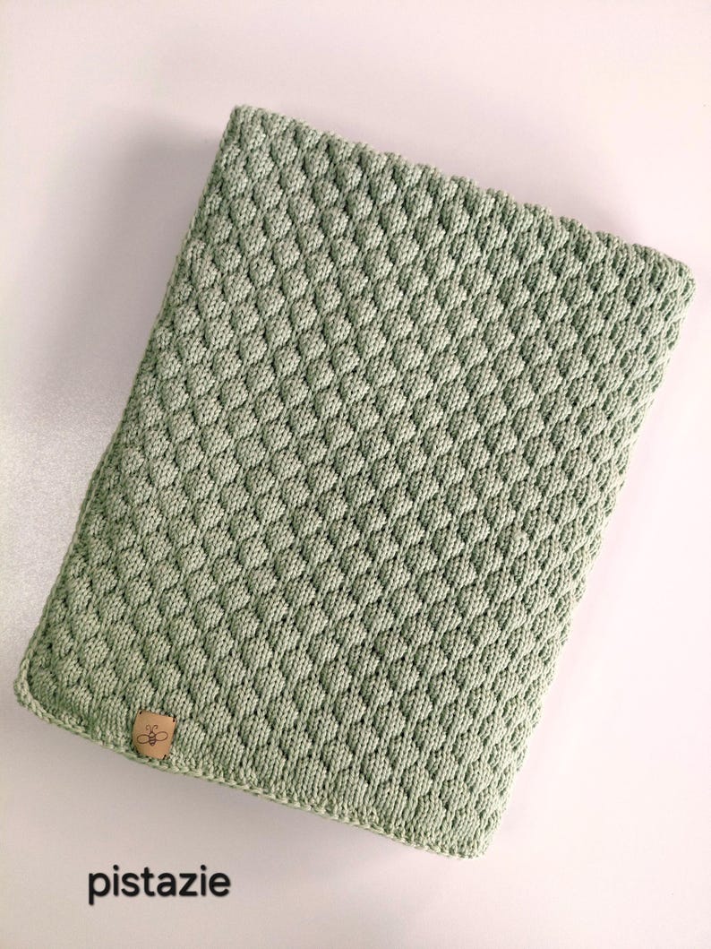 May include: A rectangular sage green knitted cover featuring a textured diamond pattern. A small tan leather-like tag with a bee icon is attached to the bottom left corner. The word "pistazie" is printed in black text below the object.