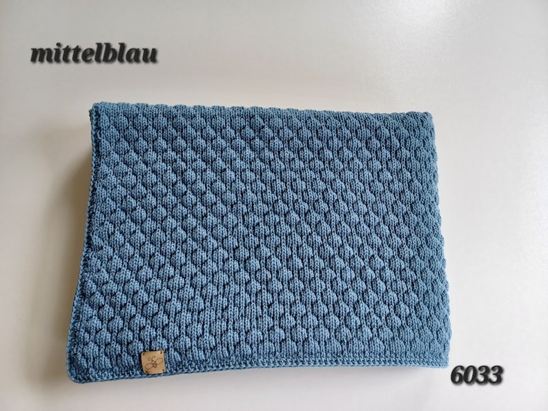 May include: A rectangular medium blue knitted blanket featuring a textured honeycomb pattern. The fabric has a small tan leather tag with a bee icon in the bottom left corner. The text "mittelblau" and "6033" are visible in the corners.