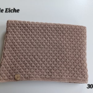 May include: A light brown knitted blanket featuring a textured diamond pattern across the surface. A small rectangular leather-like tag with a bee logo is attached to the bottom left corner. The text "helle Eiche" and "3041" appear in the image.