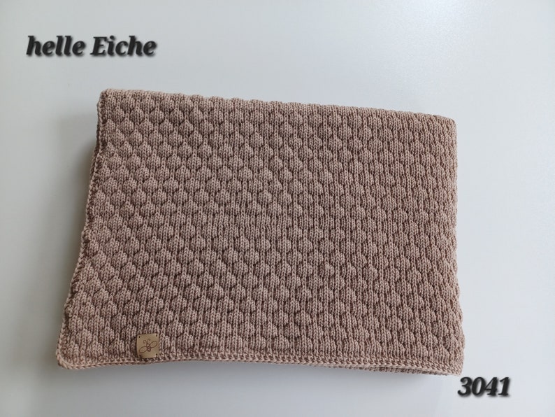 May include: A light brown knitted blanket featuring a textured diamond pattern across the surface. A small rectangular leather-like tag with a bee logo is attached to the bottom left corner. The text "helle Eiche" and "3041" appear in the image.
