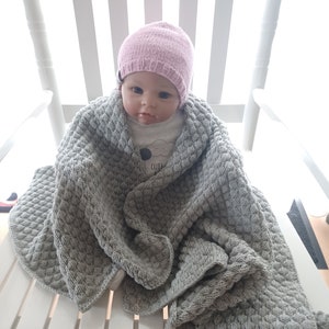 以下が含まれることがあります： ピンクのニット帽とグレーのニットブランケットを着用した赤ちゃん人形。人形は白いロッキングチェアに座っています。