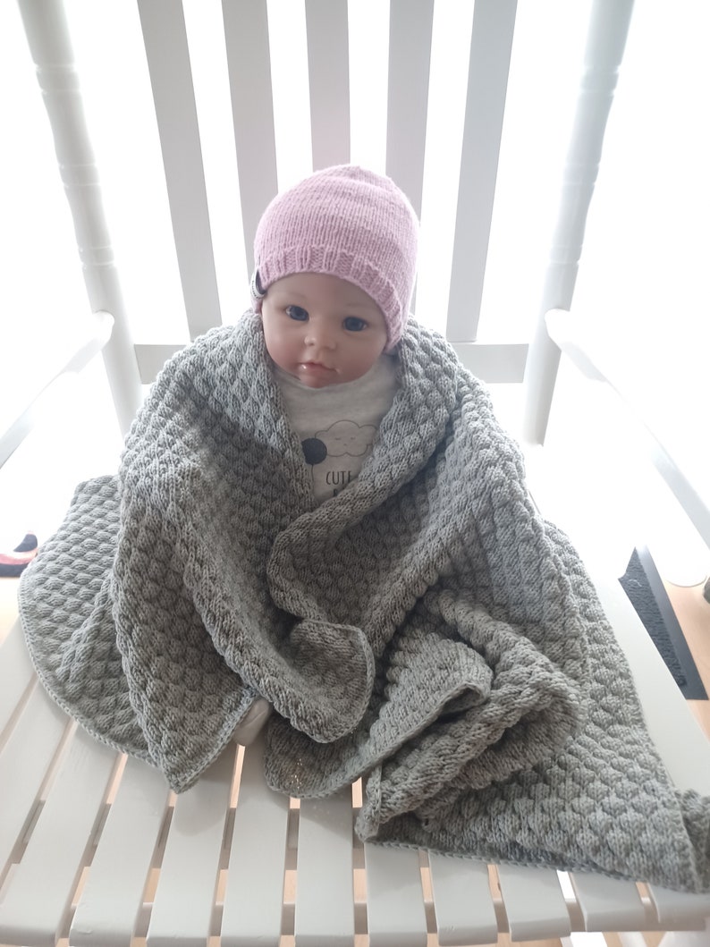 以下が含まれることがあります： ピンクのニット帽とグレーのニットブランケットを着用した赤ちゃん人形。人形は白いロッキングチェアに座っています。