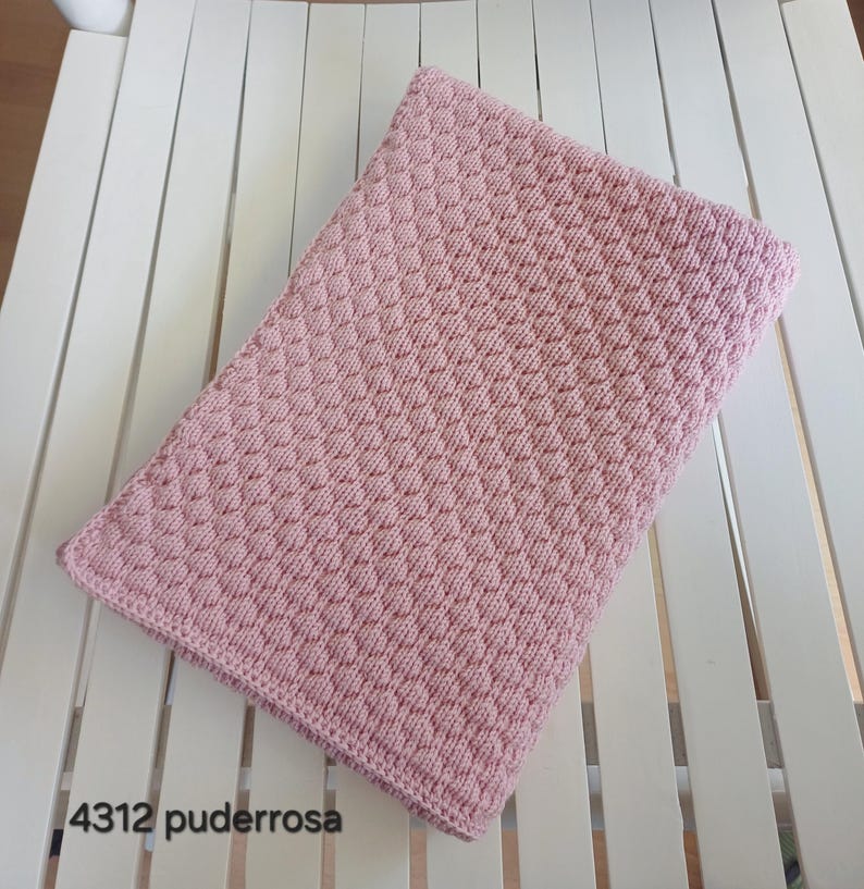 May include: A rectangular, light pink knitted blanket featuring a consistent, raised honeycomb or bubble texture pattern across the entire surface. The edges are finished with a simple, flat knit border. The text 4312 puderrosa is visible.