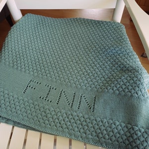 May include: A sage green knitted blanket featuring a textured diamond pattern throughout the fabric. A smooth horizontal band across the lower section displays the name FINN integrated into the knit using an openwork eyelet stitch technique.