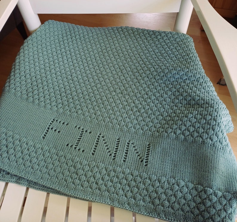 May include: A sage green knitted blanket featuring a textured diamond pattern throughout the fabric. A smooth horizontal band across the lower section displays the name FINN integrated into the knit using an openwork eyelet stitch technique.