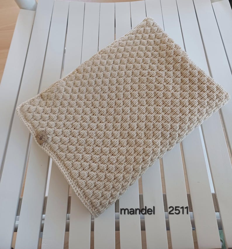 May include: A rectangular beige knitted blanket featuring a consistent textured bobble stitch pattern throughout. A small brown rectangular tag with a black logo is attached to the side. The text mandel 2511 is printed in black on the surface.