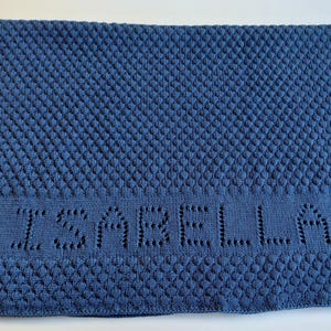 May include: A rectangular blue knit blanket featuring a textured bobble stitch pattern across the main body and borders. The center section displays the name ISABELLA integrated into the knit design using an openwork technique. A small tag is attached.