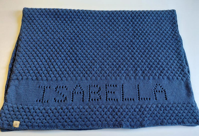 May include: A rectangular blue knit blanket featuring a textured bobble stitch pattern across the main body and borders. The center section displays the name ISABELLA integrated into the knit design using an openwork technique. A small tag is attached.