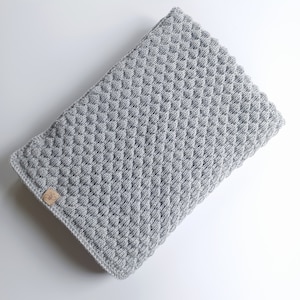 以下が含まれることがあります： 小さな茶色のラベルが付いたグレーのクロシェ編みベビーブランケット。ブランケットは、テクスチャのある隆起したパターンになっています。