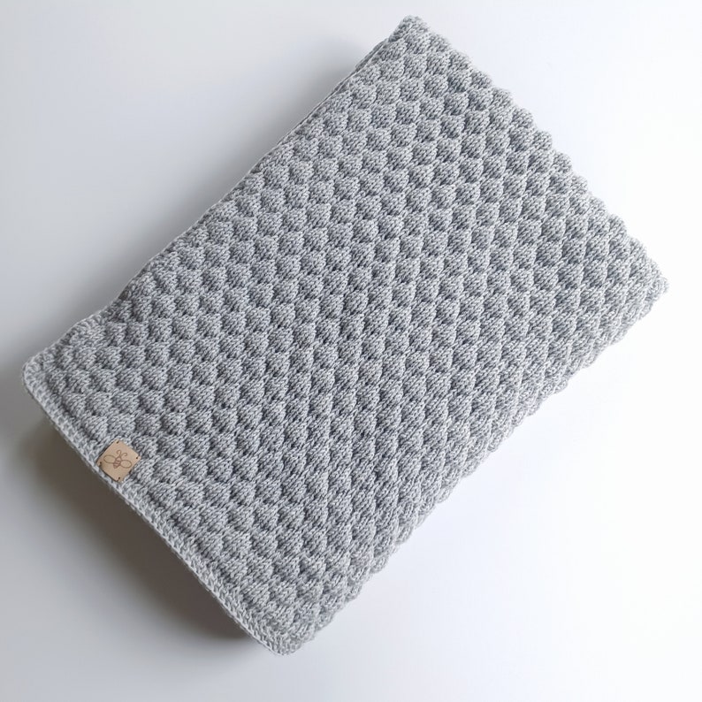 以下が含まれることがあります： 小さな茶色のラベルが付いたグレーのクロシェ編みベビーブランケット。ブランケットは、テクスチャのある隆起したパターンになっています。