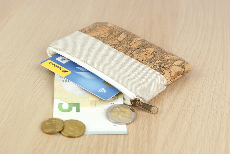 Puede incluir: Una peque&ntilde;a bolsa con cremallera de corcho y tela con un tirador de cremallera blanco. La bolsa est&aacute; abierta y contiene una tarjeta de cr&eacute;dito azul, un billete de cinco euros y dos monedas de oro.