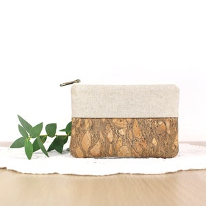 Puede incluir: Una peque&ntilde;a bolsa con cremallera con la parte superior de lino beige y la parte inferior de corcho. La bolsa est&aacute; sobre un encaje blanco sobre una superficie de madera.