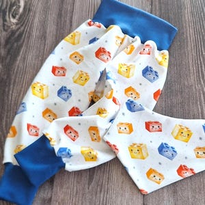 Könnte beinhalten: Weiße Babyhose und passendes Lätzchen mit farbenfrohem Blockmuster. Die Hose hat blaue Bündchen und einen blauen Bund. Die Blöcke sind rot, gelb und blau mit lächelnden Gesichtern. Das Lätzchen ist dreieckig.