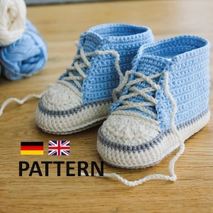 Könnte beinhalten: Ein Paar hellblaue und weiße gehäkelte Babyschuhe mit weißen Schnürsenkeln. Die Turnschuhe haben einen grauen Streifen um die Sohle und werden auf einer Holzoberfläche präsentiert. Wollknäuel in Blau- und Grautönen sind im Hintergrund. Das Wort "PATTERN" ist gedruckt.