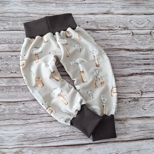 Könnte beinhalten: Hellgraue Babyhose mit einem sich wiederholenden Muster aus Aquarellwerkzeugen, darunter Hämmer, Schraubenschlüssel und Zangen. Der Bund und die Bündchen sind dunkelbraun. Die Hose ist aus weichem Stoff.