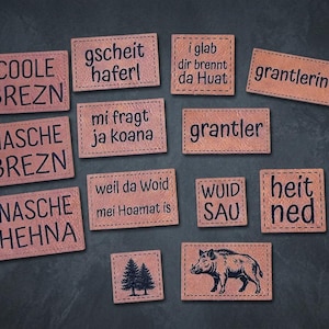 Könnte beinhalten: Eine Sammlung rechteckiger, hellbrauner Lederaufnäher mit schwarzer Schrift und Illustrationen. Die Texte umfassen COOLE BREZN, NASCHE BREZN, NASCHE HEHNA, gscheit haferl, i glab dir brennt da Huat, mi fragt ja koana, grantlerin, grantler, weil da Woid mei Hoamat is, WUID SAU und heit ned. Ein Aufnäher zeigt einen Tannenbaum, ein anderer ein Wildschwein.
