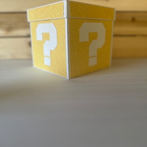 May include: A yellow box with a glittery finish and white question marks on each side. The box is a popular theme for party decorations and games.