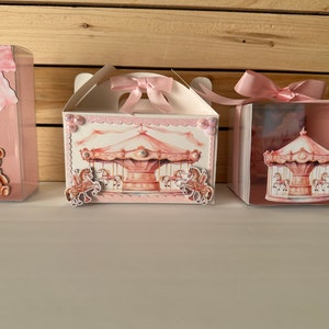 Puede incluir: Tres cajas de regalo rosas y blancas con temática de carrusel. Las cajas están decoradas con cintas rosas y un caballo de carrusel. Una caja tiene un oso de peluche dentro.