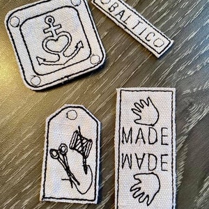 Puede incluir: Cuatro parches de tela blanca con bordados negros. Un parche presenta un ancla con un corazón, otro dice "BALTICO", un tercero dice "MADE" y "WADE" con una huella de mano arriba y abajo de las palabras, y el último parche presenta un par de tijeras y una bobina de hilo.